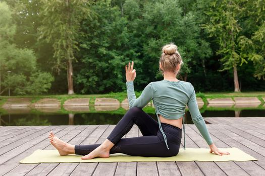 A woman in a green top and leggings, sits on a wooden platform by a pond in a park in summer, does yoga, performing a table pose. Copy space. Back view