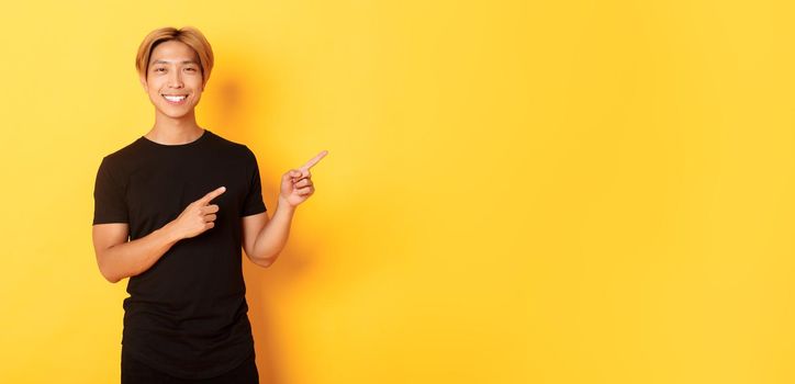 Portrait of satisfied smiling blond guy, asian student pointing fingers right, showing way, standing yellow background.