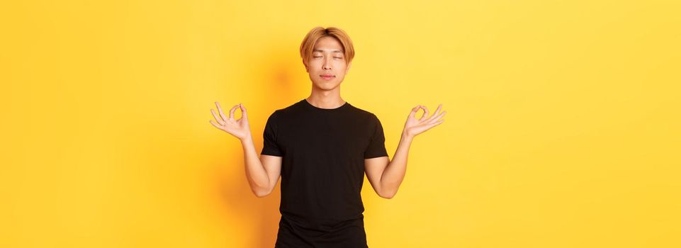 Portrait of relaxed and peaceful handsome asian man with blond hair, close eyes and meditating, attend yoga, standing yellow background.
