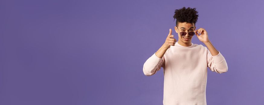 Portrait of cheeky handsome young man wearing sunglasses, take them off and show thumb-up, like or approval, recommendation sign, smirk satisfied, saying all good, alright, purple background.