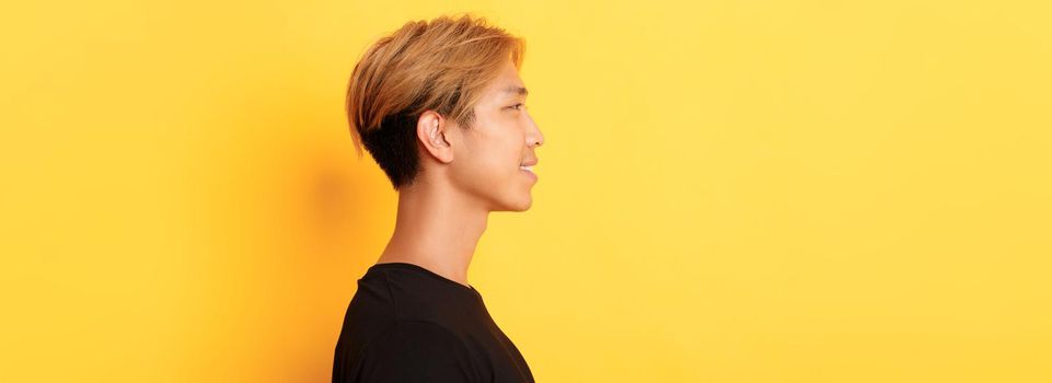 Profile of stylish handsome asian guy with fair hair looking left and smiling, standing over yellow background.