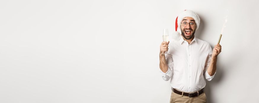 Winter holidays and celebration. Handsome bearded man having New Year party, holding firework sparkler and champagne, wearing santa hat, white background.