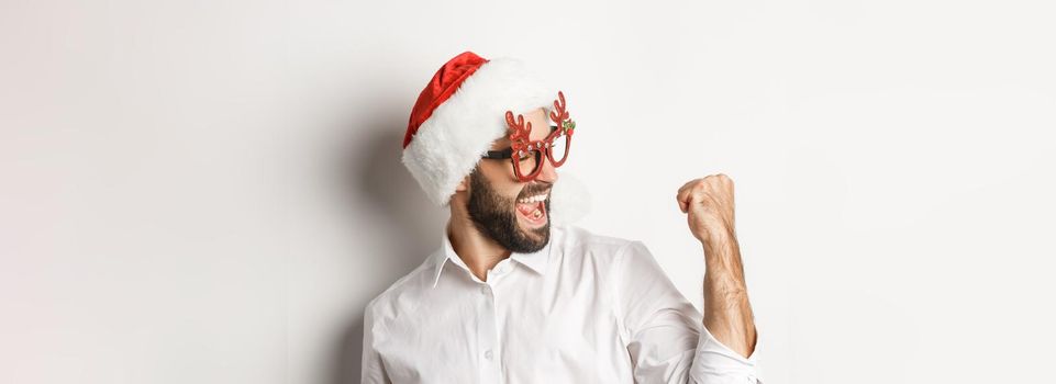 Close-up of happy bearded man wearing santa hat and christmas party glasses, making fist pump and rejoicing, achieve goal and celebrating, white background.