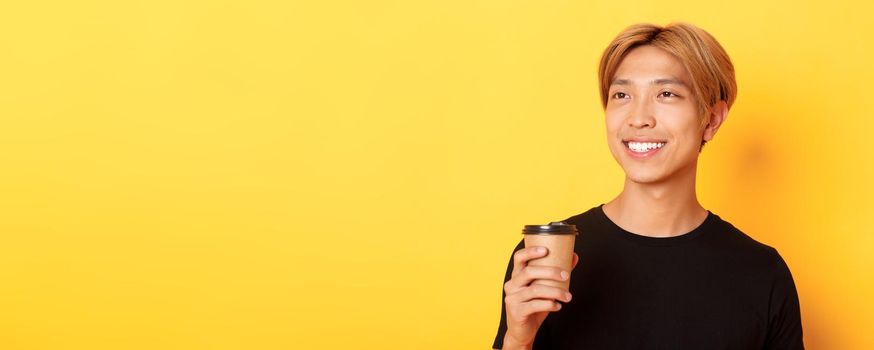 Close-up of relaxed and happy stylish asian blond guy, smiling and looking left dreamy while drinking coffee from takeaway cup, standing over yellow background.