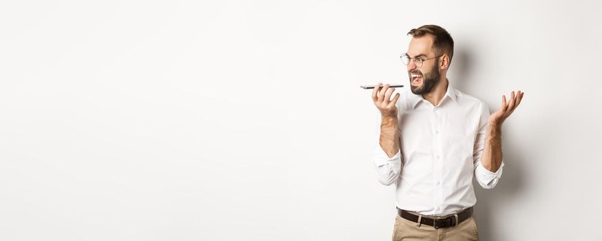 Angry business man shouting at speakerphone, record voice message in mad state, standing against white background.