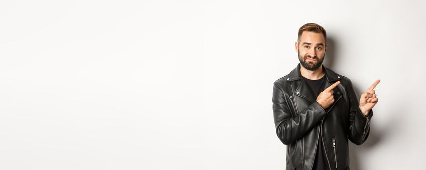 Skeptical and doubtful guy in black leather jacket, shrugging while pointing at upper right corner promo offer, standing over white background.