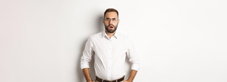 Shocked and displeased businessman in glasses, gasping and looking upset at camera, standing over white background. Copy space