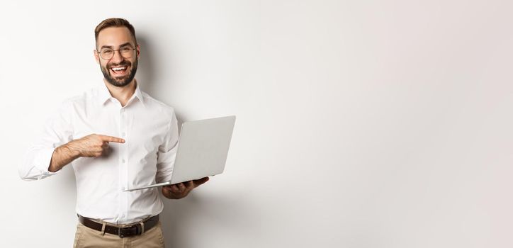 Business. Handsome manager in glasses working on laptop, pointing at computer and smiling pleased, standing over white background.