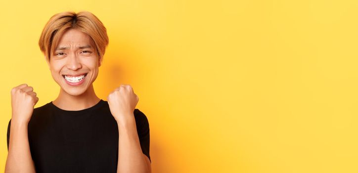 Close-up of lucky happy asian guy winning prize, making fist pump gesture and smiling satisfied, triumphing over achievement, standing yellow background.