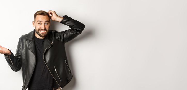 Confused bearded guy in cool leather jacket shrugging, scratch head puzzled and clueless, standing over white background.