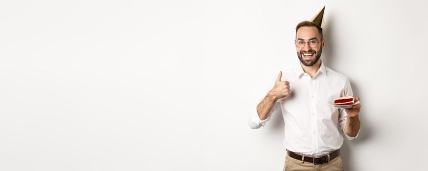 Holidays and celebration. Satisfied man enjoying b-day party, holding birthday cake and showing thumb up in approval, recommending something, white background.