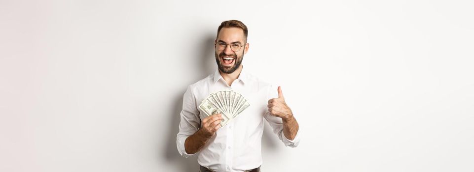 Excited rich man holding money, showing thumb up in approval, standing over white background. Copy space
