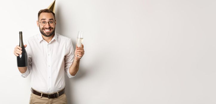 Celebration and holidays. Happy birthday guy enjoying b-day party, wearing funny cone hat and drinking champagne, white background.