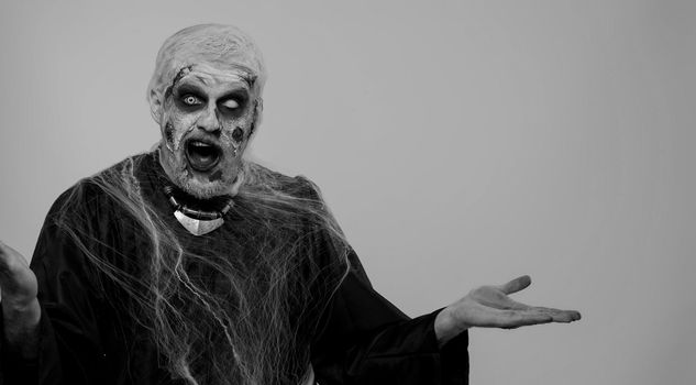Creepy man with bloody scars face, Halloween stylish zombie make-up. Scary wounded undead guy showing thumbs up and pointing at left on blank space. Place for your advertisement logo. Sinister monster