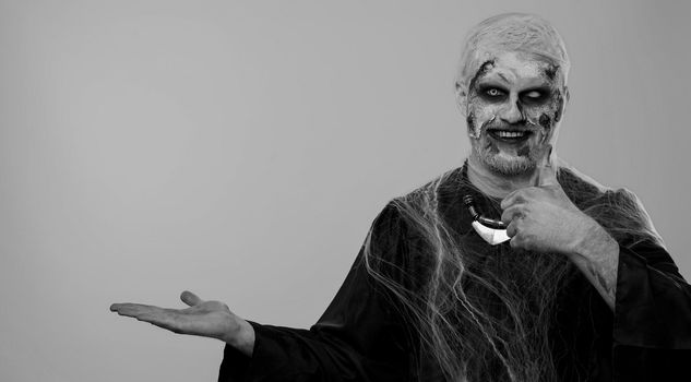 Creepy man with bloody scars face, Halloween stylish zombie make-up. Scary wounded undead guy showing thumbs up and pointing at right on blank space. Place for advertisement logo. Sinister monster