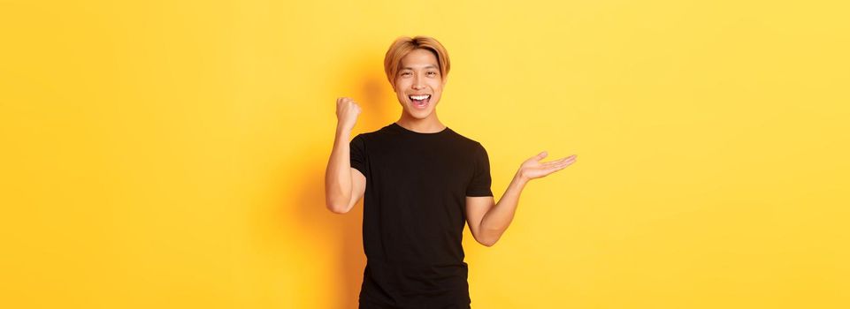 Satisfied and happy attractive korean guy smiling, fist pump with rejoice, holding something on hand, standing yellow background.