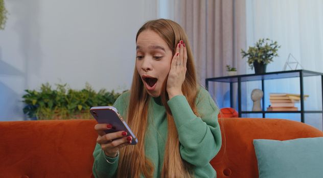Oh my God Wow. Excited happy joyful winner girl use smartphone typing browsing found out great win good news celebrate victory. Young child kid with mobile phone sits at home in room on orange couch