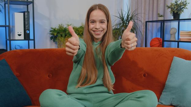 Happy teen girl shouting, celebrating success, winning, goal achievement, good news, raising fists in gesture I did it, lottery luck at home apartment. Young child kid indoors in living room on couch
