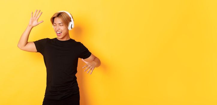 Happy attractive asian guy with blond hair, singing along and dancing as listening to music in wireless headphones, standing yellow background.