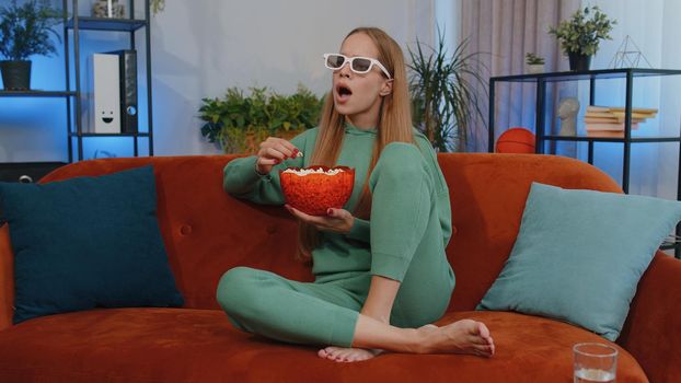 Excited teen girl sitting on sofa eating popcorn and watching interesting tv serial, sport game, film, online social media movie content online at home. Young child kid enjoying domestic entertainment