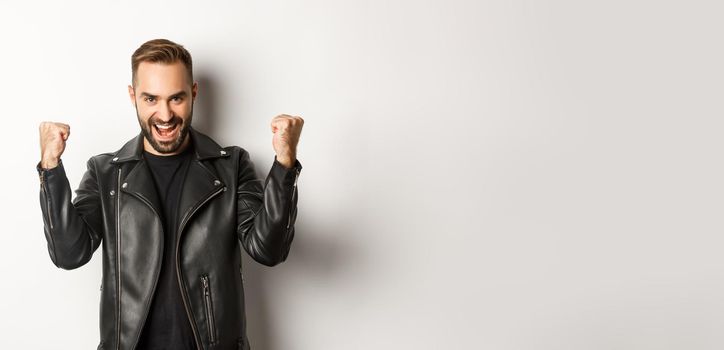 Confident and assertive man in leather jacket making fist pump, rejoicing of winning, feeling encouraged and satisfied, white background.