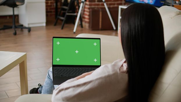 Woman sitting on sofa at home while looking at laptop having green screen chroma key isolated display. Young adult person sitting on couch inside apartment with portable computer on lap.