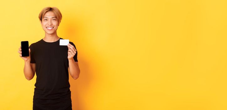 Portrait of handsome young asian guy showing smartphone screen, banking app and credit card, standing yellow background and smiling.