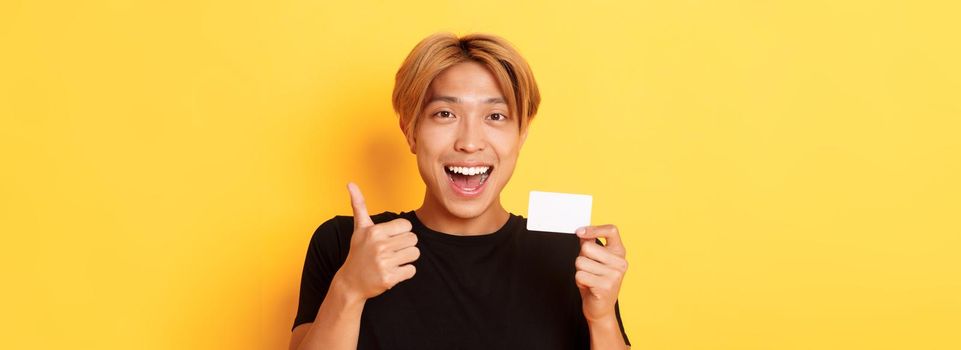 Close-up of happy and satisfied asian handsome guy, showing credit card and thumbs-up in approval, smiling amazed, standing yellow background.