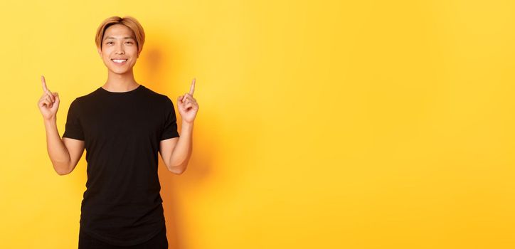 Portrait of handsome smiling asian guy in black t-shirt pointing fingers up and looking satisfied, yellow background.
