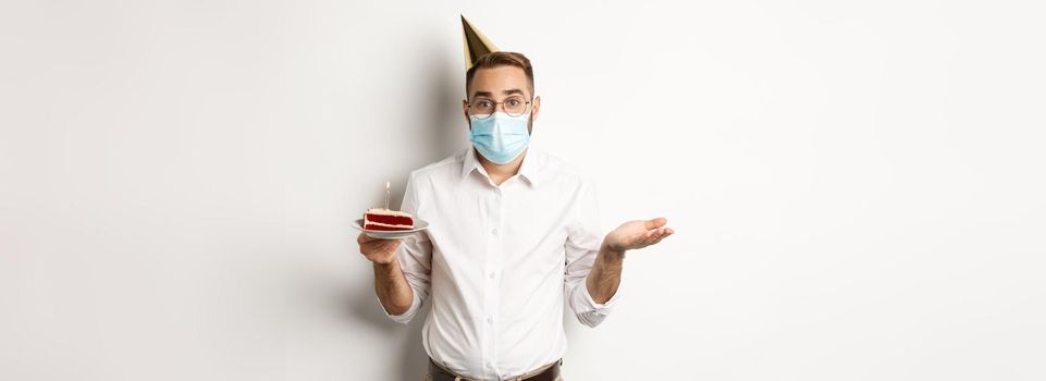 Coronavirus, quarantine and holidays. Confused man in face mask, holding birthday cake and shrugging, standing over white background clueless.