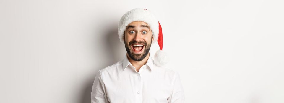 Close-up of happy bearded guy in santa hat, looking surprised, celebrating christmas, standing over white background.