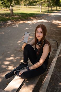 Young fit woman in sportswear and headphones sitting on bench in park and demonstrating mobile phone with blank screen