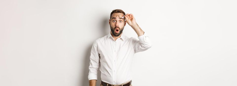 Surprised businessman taking-off his glasses, looking with amazement at camera, standing over white background.