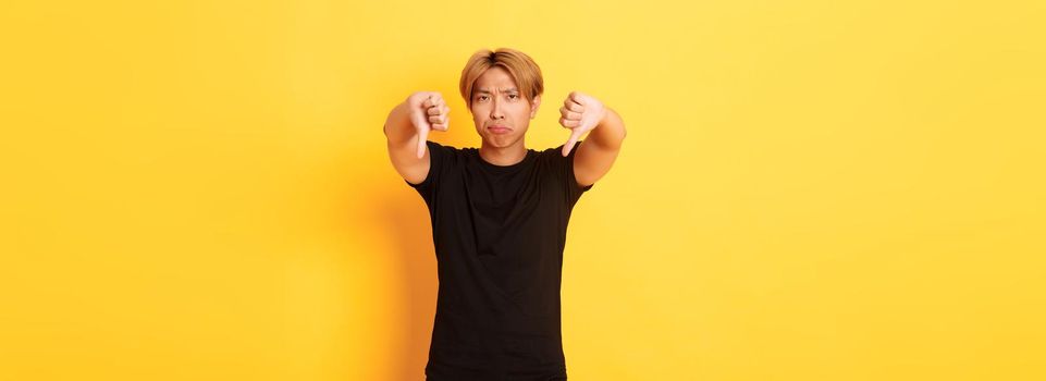 Portrait of disappointed and upset asian man showing thumbs-down, grimacing displeased, yellow background.