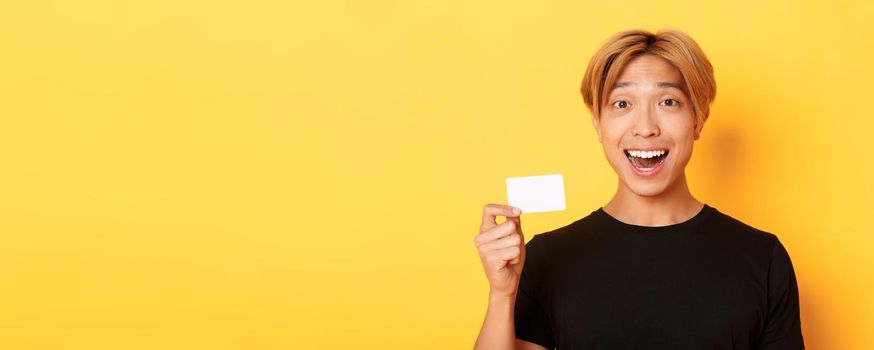 Close-up of excited surprised handsome asian guy, smiling amazed and showing credit card, standing yellow background.