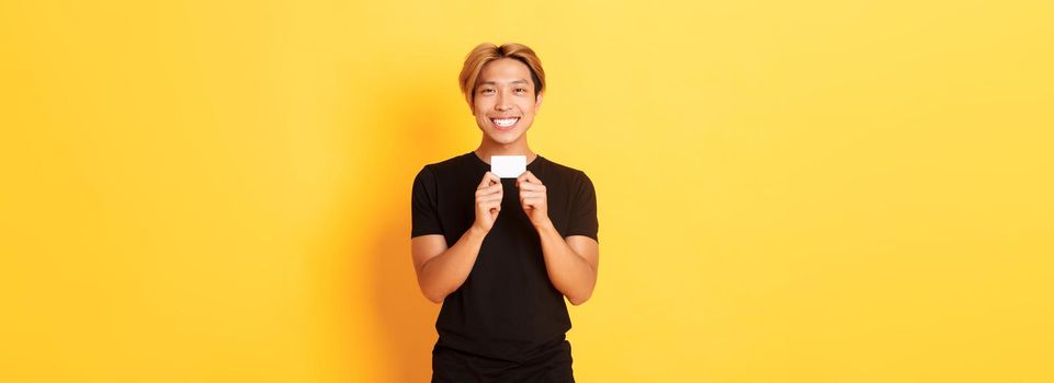 Excited happy asian guy smiling and showing credit card, standing yellow background.
