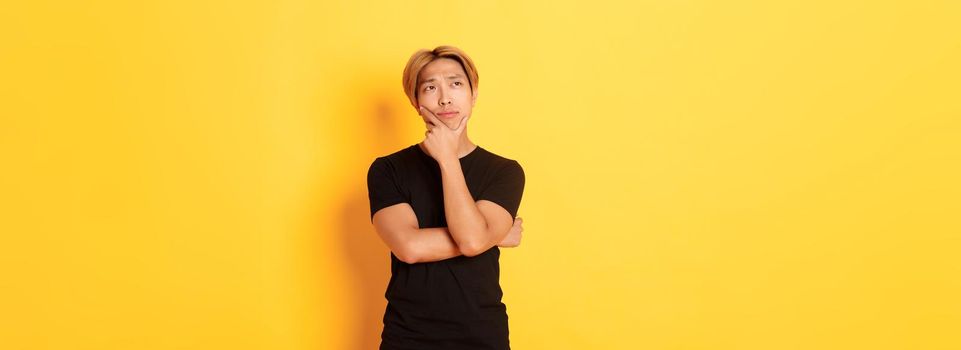 Portrait of thoughtful attractive guy in black t-shirt looking upper left corner. Young man pondering idea, standing yellow background.