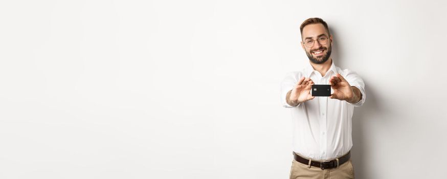 Handsome man in glasses holding a credit card, smiling pleased, standing over white background.