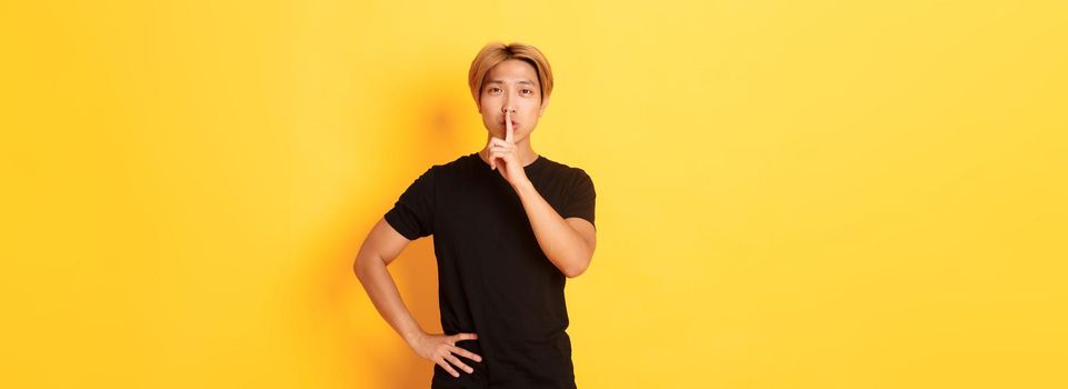 Portrait of attractive blond asian man shushing, press finger to lips and hush, yellow background.