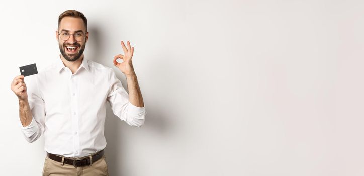 Handsome man showing his credit card and okay sign, recommending bank, standing over white background. Copy space