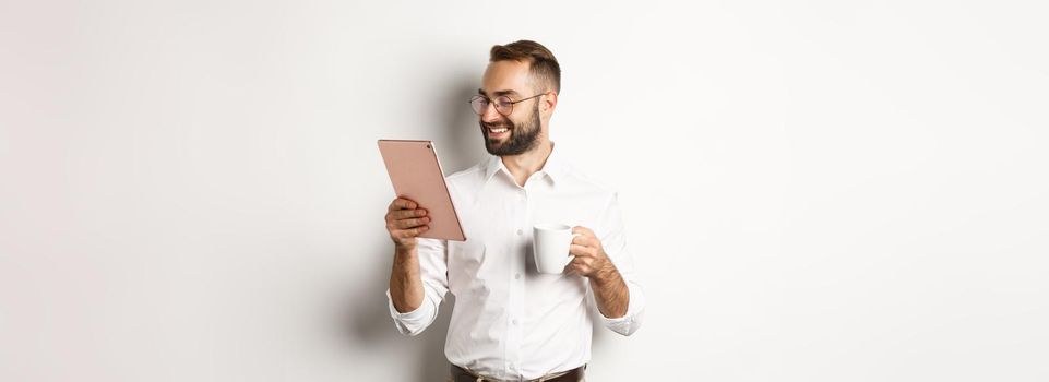 Handsome businessman drinking coffee and reading on digital tablet, smiling pleased, standing over white background.