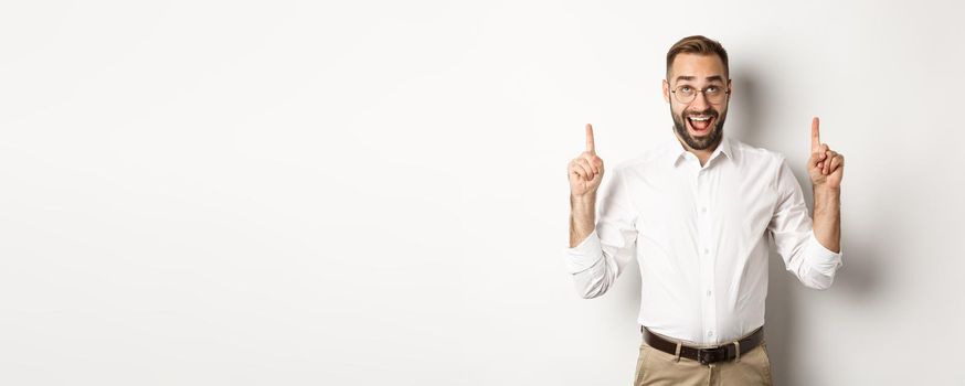 Excited businessman checking out advertisement, pointing and looking up with happy face, standing against white background.