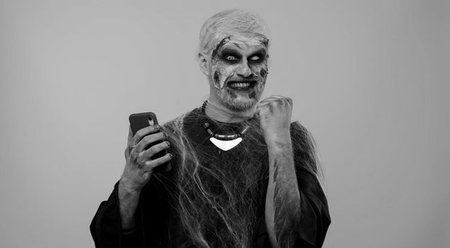 Happy excited sinister man in carnival costume of Halloween crazy zombie looking smartphone display sincerely rejoicing win, receiving message with good news, success luck. Cosplay wounded undead