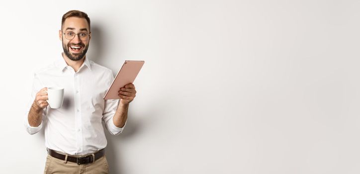 Excited manager reading on digital tablet, working and drinking coffee, standing against white background.