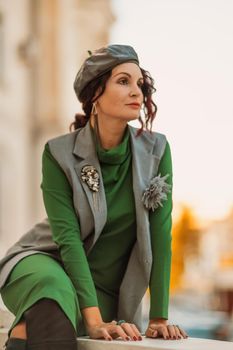 Outdoor fashion portrait of an elegant fashionable brunette model in a beret, green dress and a gray waistcoat posing at sunset in a european city in autumn