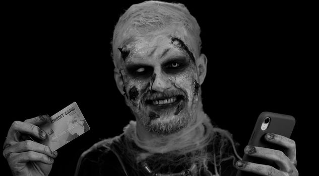 Sinister man with horrible scary Halloween zombie make-up using credit bank card and mobile phone, transferring money, purchases online shopping. Dead guy with wounded bloody scars face, black room