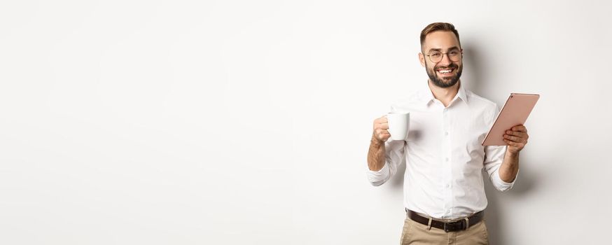 Satisfied boss drinking tea and using digital tablet, reading or working, standing over white background.
