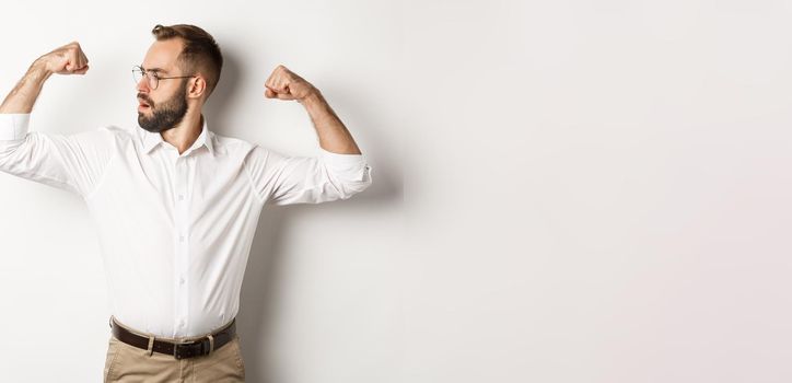 Successful businessman flex biceps, showing muscles and looking confident, feeling strong, standing over white background.