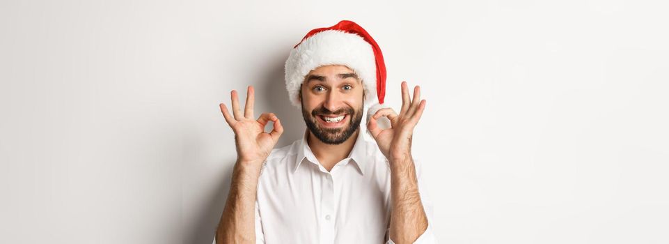 Party, winter holidays and celebration concept. Joyful man enjoying christmas and showing okay sign, smiling satisfied, wearing santa hat, white background.