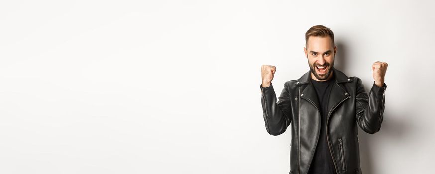 Confident and assertive man in leather jacket making fist pump, rejoicing of winning, feeling encouraged and satisfied, white background.